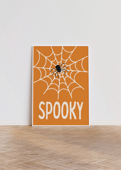 ‘Spooky’ Halloween poster featuring a spiderweb and spider on an orange background in a white frame, leaning against a white wall.