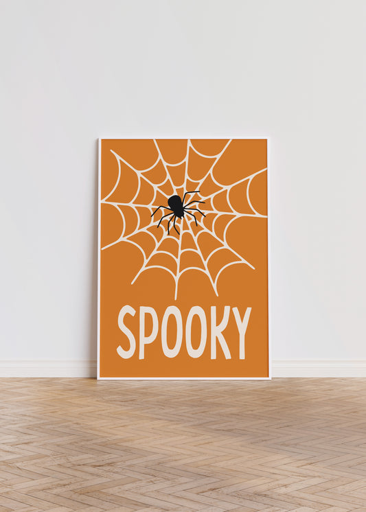 ‘Spooky’ Halloween poster featuring a spiderweb and spider on an orange background in a white frame, leaning against a white wall.
